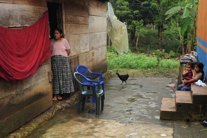 An indigenous Mayan woman and 2 children stand outside their home with a rooster nearby, in rural Guatemala.