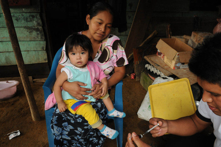 An indigenous Mayan woman holds her 1-year-old daughter as a volunteer health worker prepares a syringe with measles vaccine in rural Guatemala.