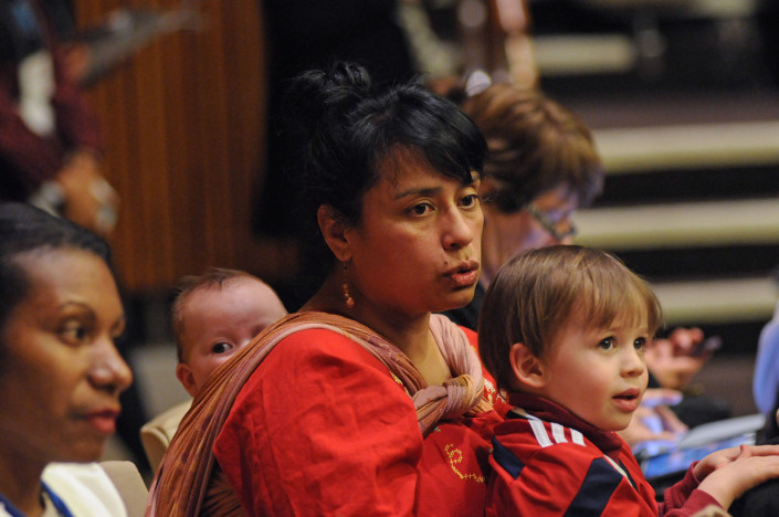 A woman carrying a baby in a sling on her back and a child on her lap, listens to speakers at a UN conference.
