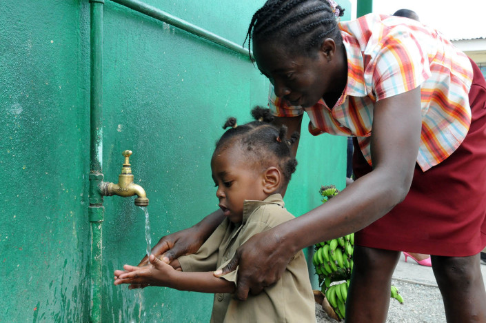 A teacher helps a toddler wash his hands at an outdoor water point outside a school in the parish of Kingston and St. Andrew, Jamaica.