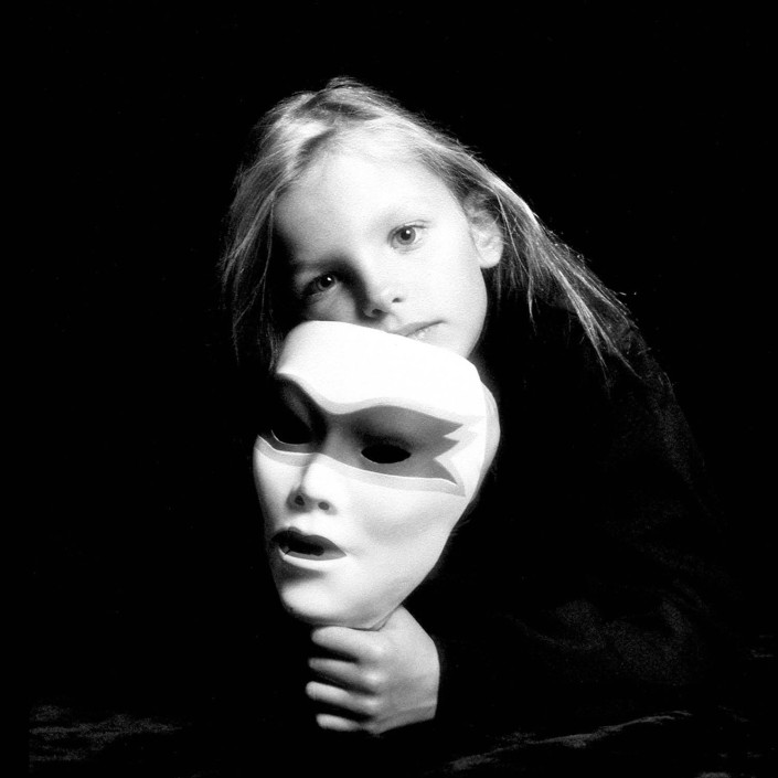A 7-year-old girl holds a white mask mirroring her features in a studio portrait.