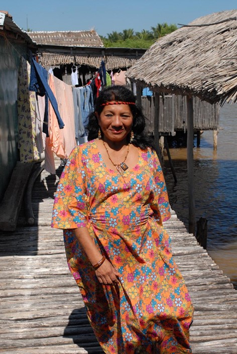 An indigenous woman advocate wearing a colorful dress, stands outside a typical Anu dwelling above a lagoon in Venezuela.