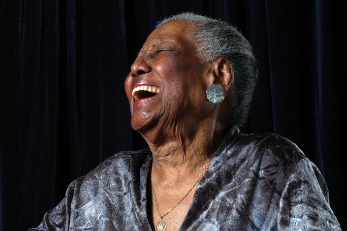 An 86-year-old elegantly-dressed woman, smiles in a hearty laugh with her head thrown back.