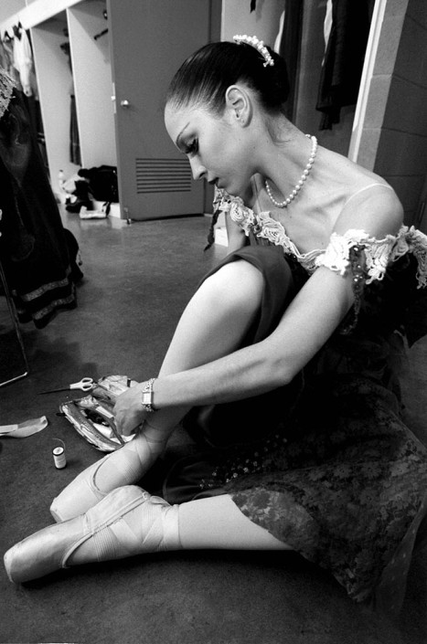 A professional ballerina dressed in lace and pearls, mends her ballet shoes before a performance.