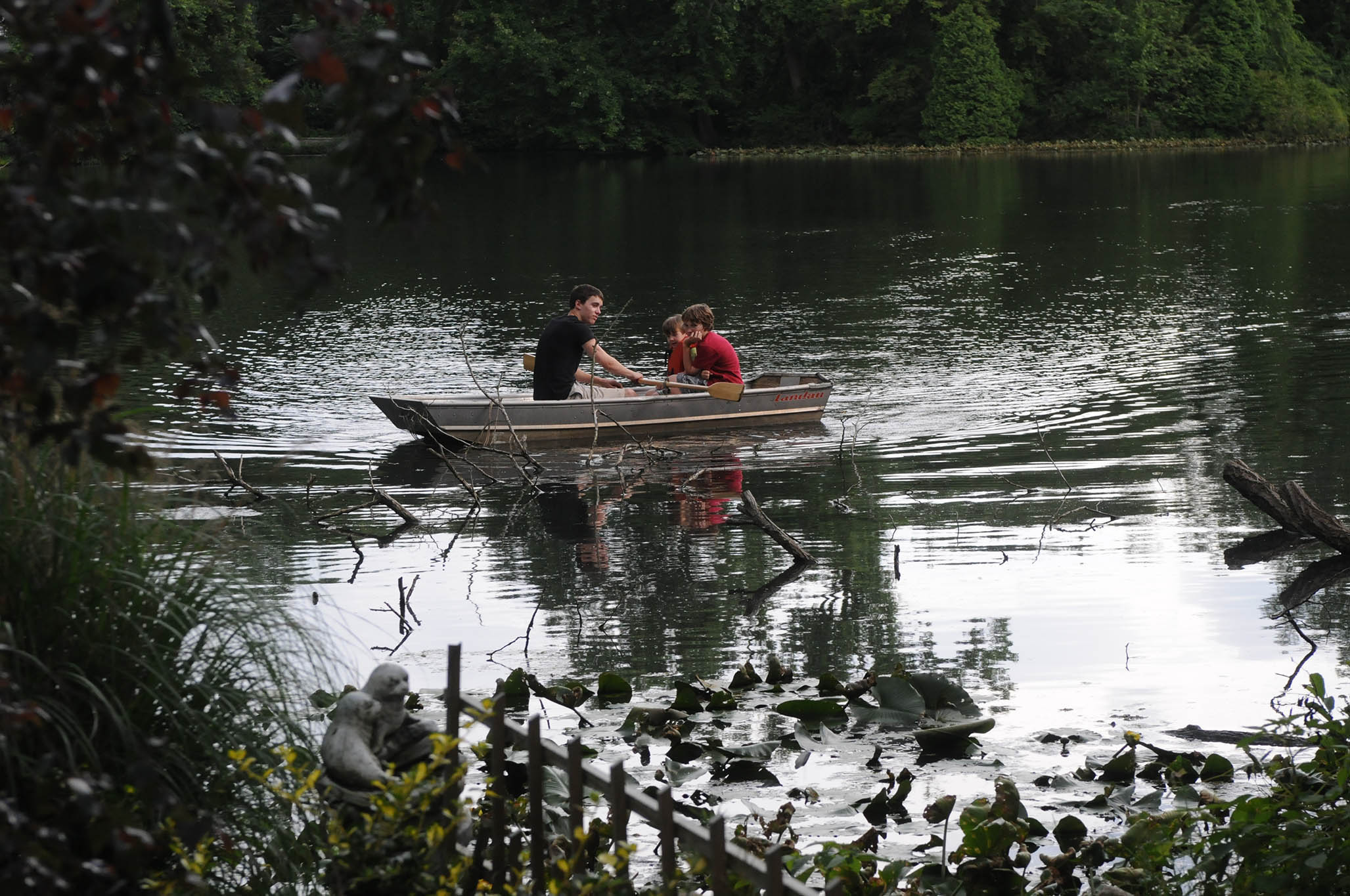 Three young boys in a rowboat on a small pond surrounded by trees and foliage.