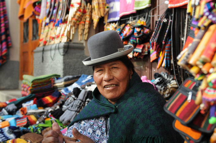 An indigenous woman wearing a bowler hat is surrounded by colorful textiles in La Paz, Bolivia.