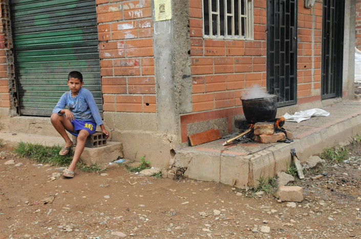 A boy sits on a cement block around the corner from a large pot cooking on a fire, in a poor neighborhood in Medellin, Colombia.
