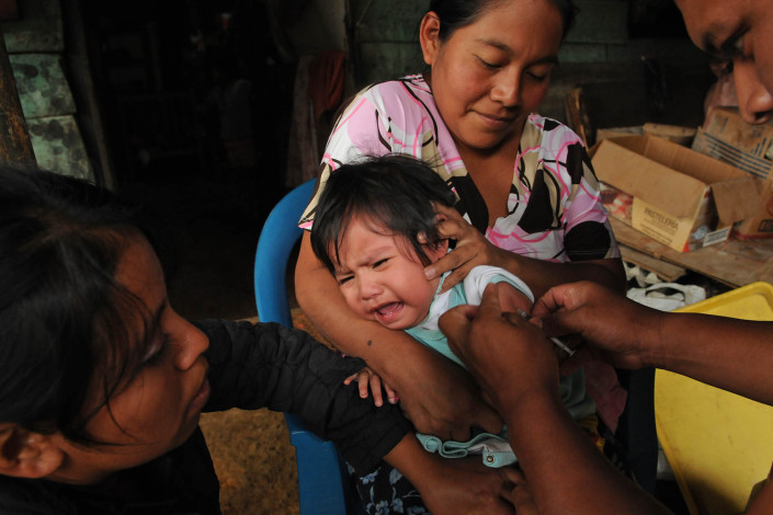 An indigenous Mayan woman holds her 1-year-old crying daughter as a volunteer health worker administers a measles vaccination in rural Guatemala.