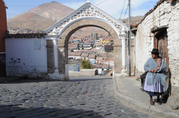 A woman stands on a sidewalk in the City of Potosí, Bolivia. Cerro Rico Mines are behind her.