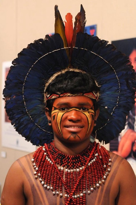 Portrait of an indigenous adolescent boy wearing traditional dress including a headdress with feathers, face paint and a large red and black beaded necklace.