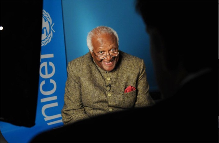 Archbishop Desmond Tutu, smiling, is interviewed in a studio with a UNICEF logo in the background.