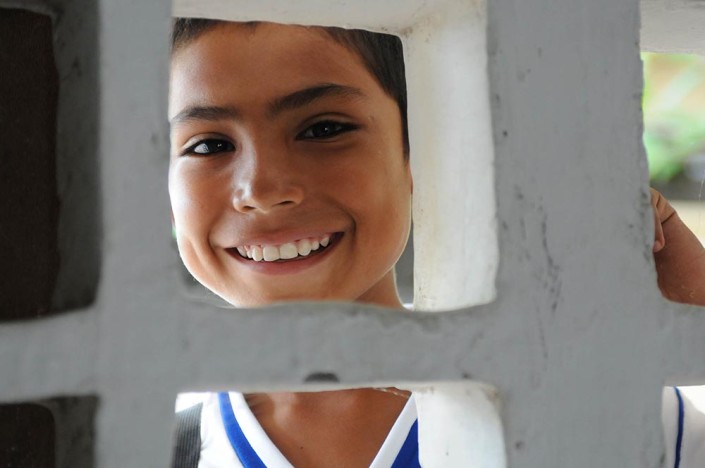 A smiling boy boy looks through a white cement block window into his classroom in Colombia.