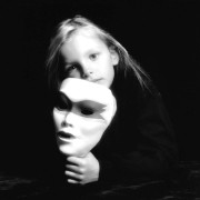 A pensive 7-year-old girl holds a mask in her hand, mirroring the look on her face, in a studio portrait.