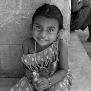 A 4-year-old smiling indigenous girl wearing a string of beads, smiles while sitting outside her home.