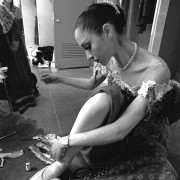 A ballerina dressed in pearls and a lace costume, mends her ballet shoes with a needle and thread before a performance.