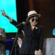 Yoko Ono, wearing a black and white fedora and black sunglasses, raises her hand in a peace sign as Hugh Jackman stands beside her.