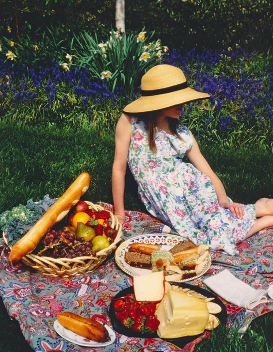 A girl in a garden, wearing a straw hat and floral dress sits on a picnic blanket filled with fresh breads, cheeses and fruits, for an illustration.