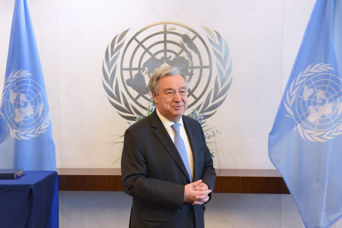 UN Secretary General Antonio Guterres stands in his conference room in front of the UN logo and between UN flags