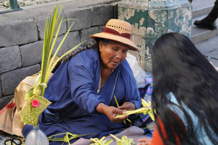 A woman wearing a straw hat sells decorative palm fronts in the shape of crosses on a street in La Paz, Bolivia.